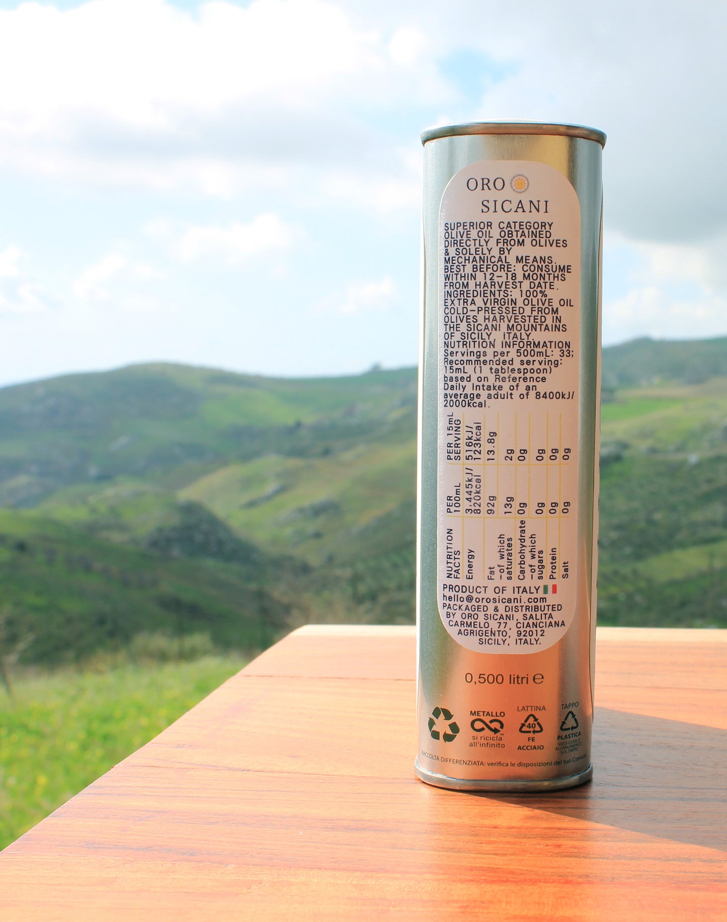 Close-up side profile of nutrition information of ORO SICANI Extra Virgin Olive Oil tin, on top of wooden table with lush, green mountains in the background.