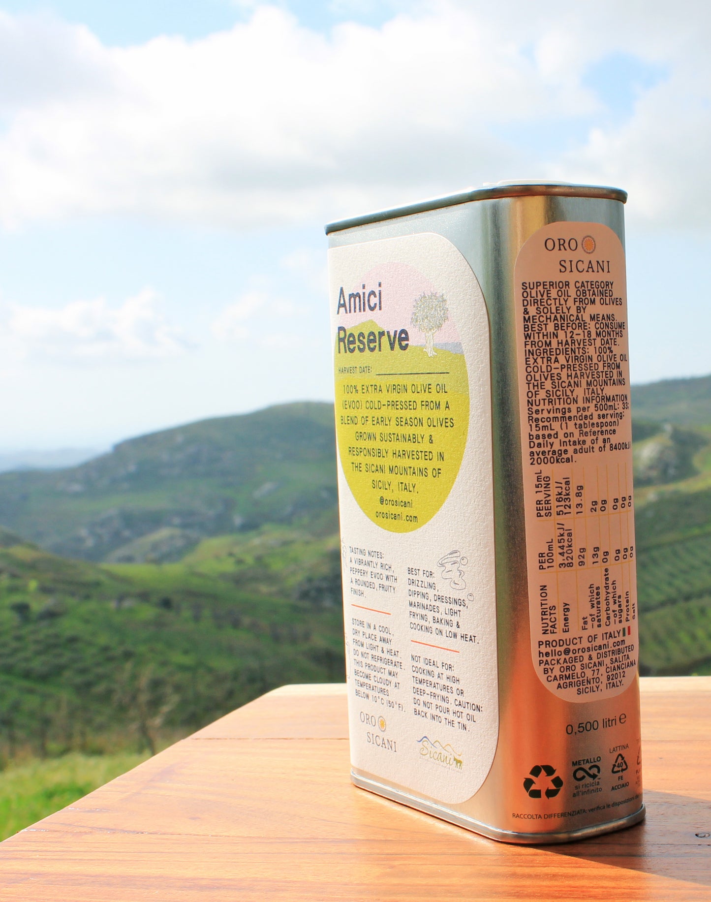 Close-up side profile of ORO SICANI Extra Virgin Olive Oil tin, on top of wooden table with lush, green mountains in the background.