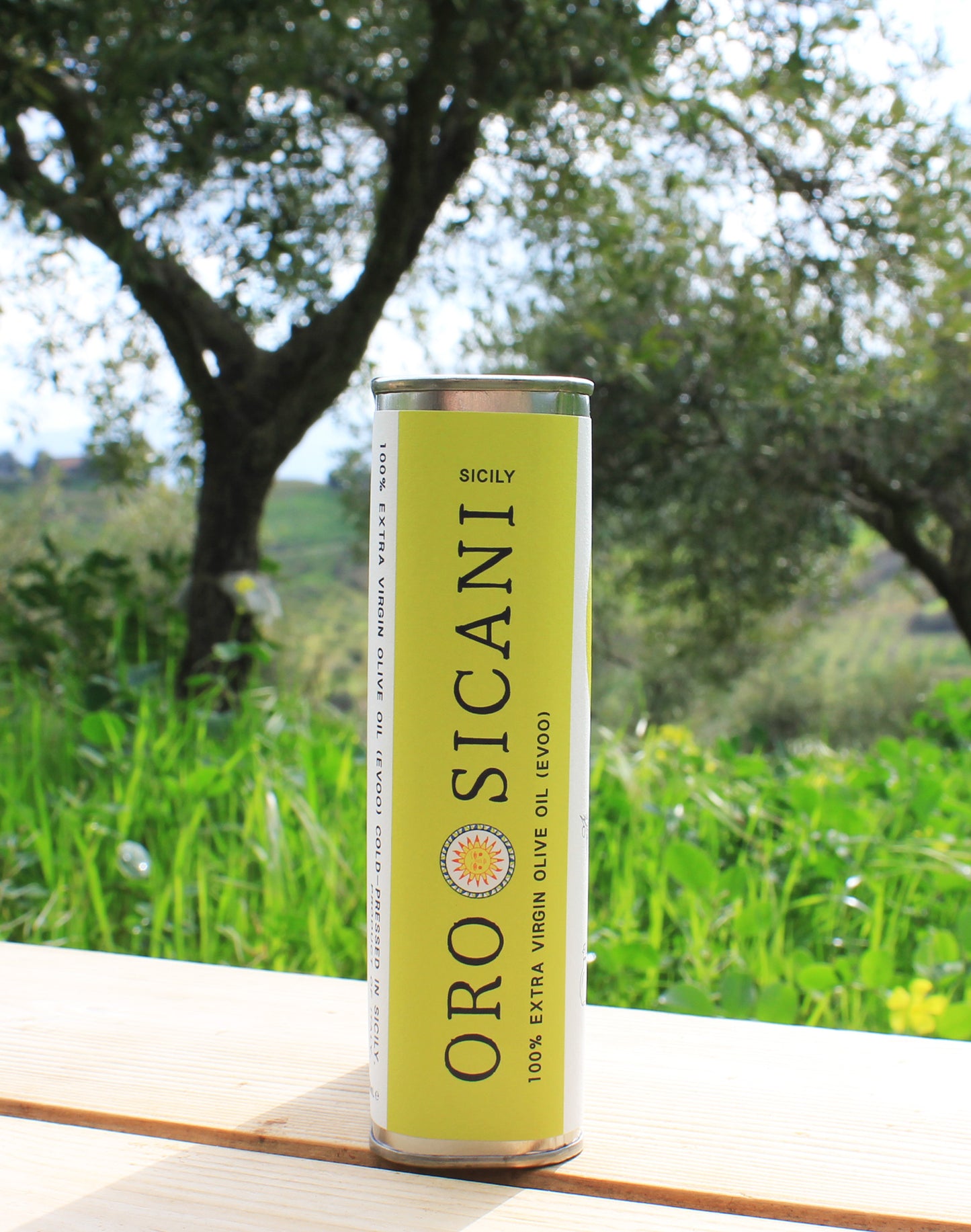 Side profile close-up of ORO SICANI Extra Virgin Olive Oil tin on top of wooden table with olive trees on ORO SICANI Estate in the background.