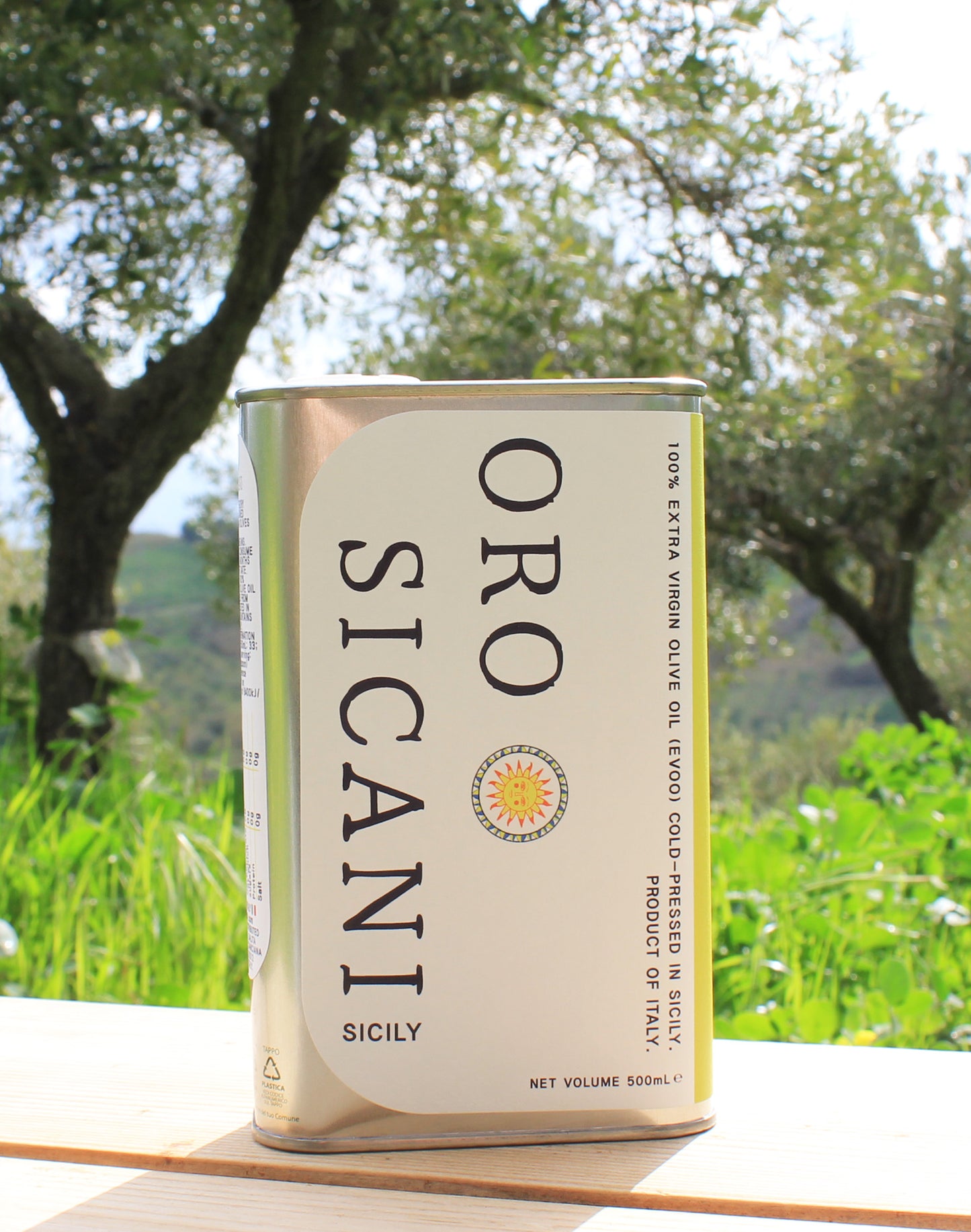 Close-up of ORO SICANI Extra Virgin Olive Oil tin on top of wooden table with olive trees on ORO SICANI Estate in the background.