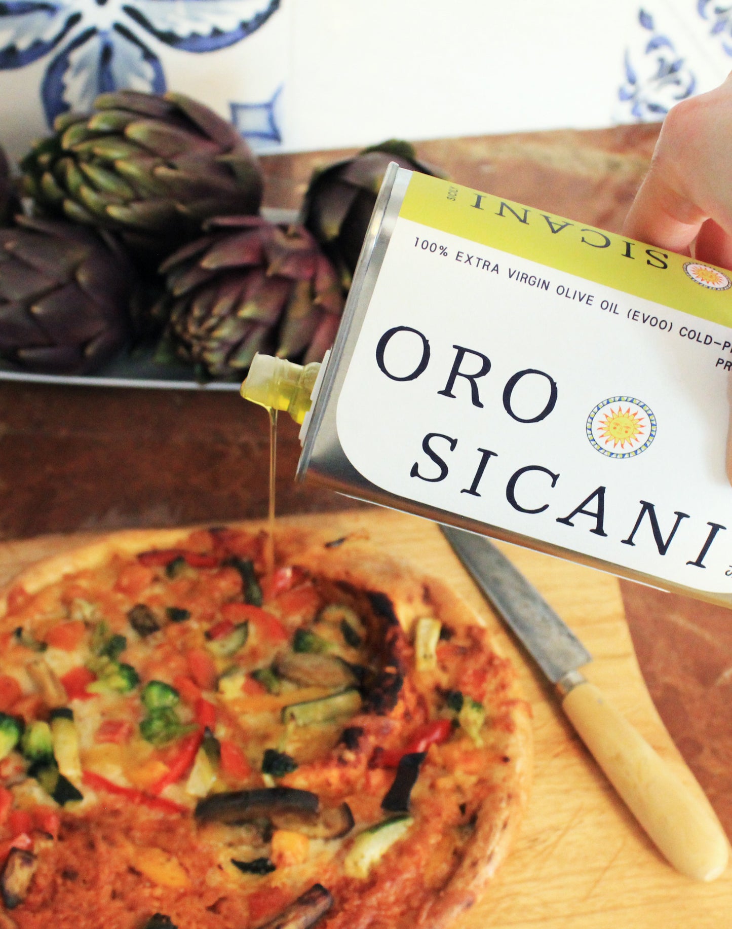 ORO SICANI Extra Virgin Olive Oil being poured over a vegetable pizza on a chopping board that's on a red marble kitchen counter with a small platter of artichokes in the background.