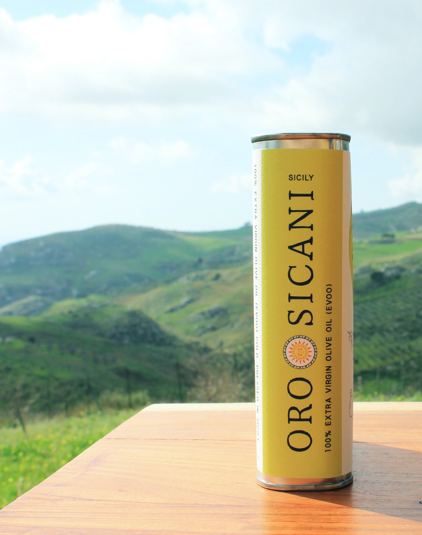 Close-up side profile of ORO SICANI Extra Virgin Olive Oil tin, on top of wooden table with lush, green mountains in the background.