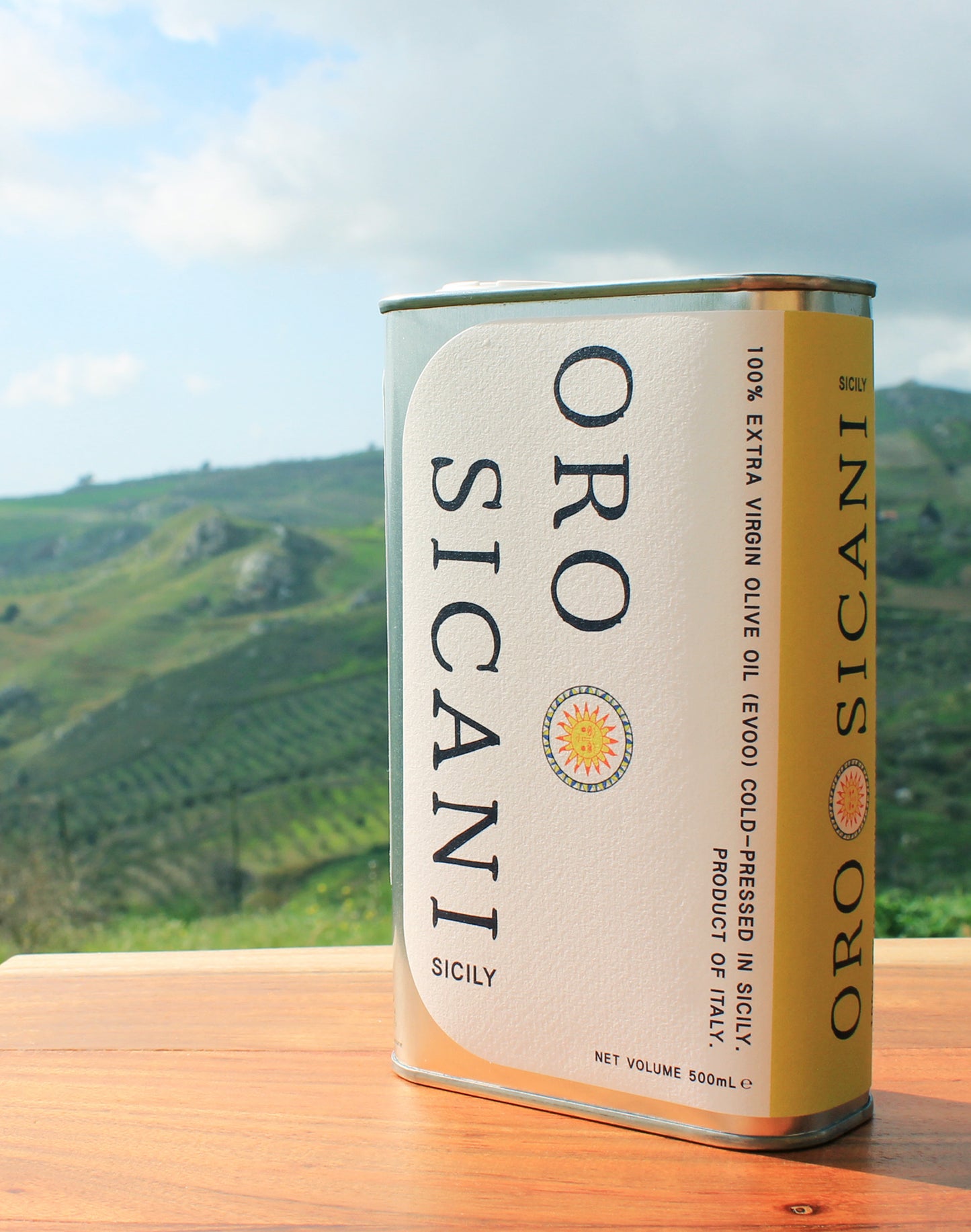 Close-up of ORO SICANI Extra Virgin Olive Oil tin, on top of wooden table with lush, green mountains in the background.