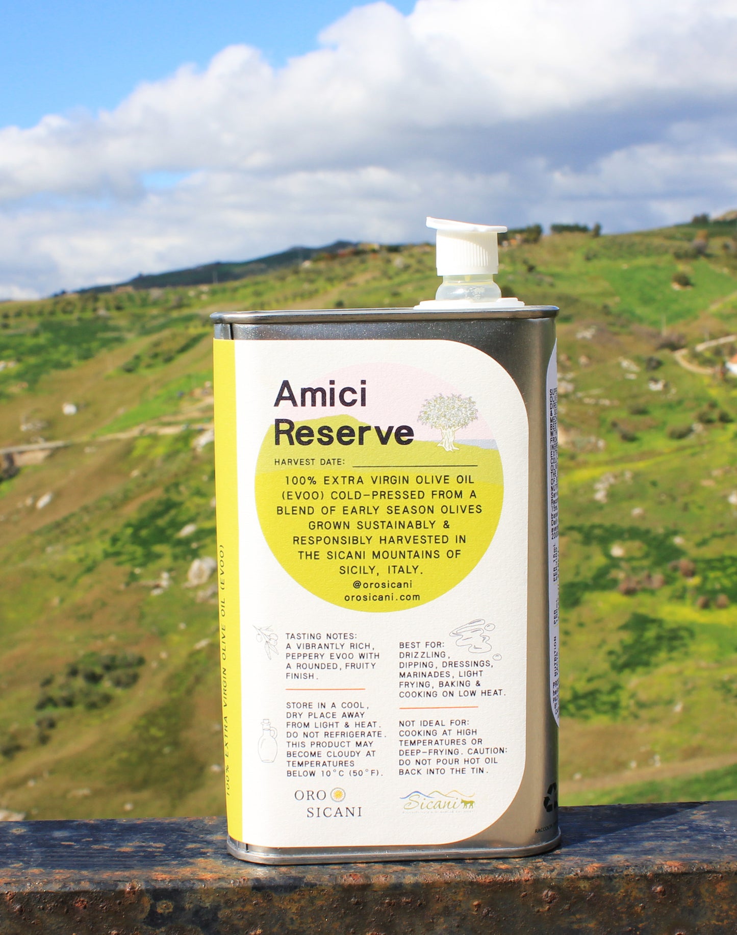 Close-up of ORO SICANI Amici Reserve Extra Virgin Olive Oil tin, placed on top of rusted railing with a lush, green mountain in the background.