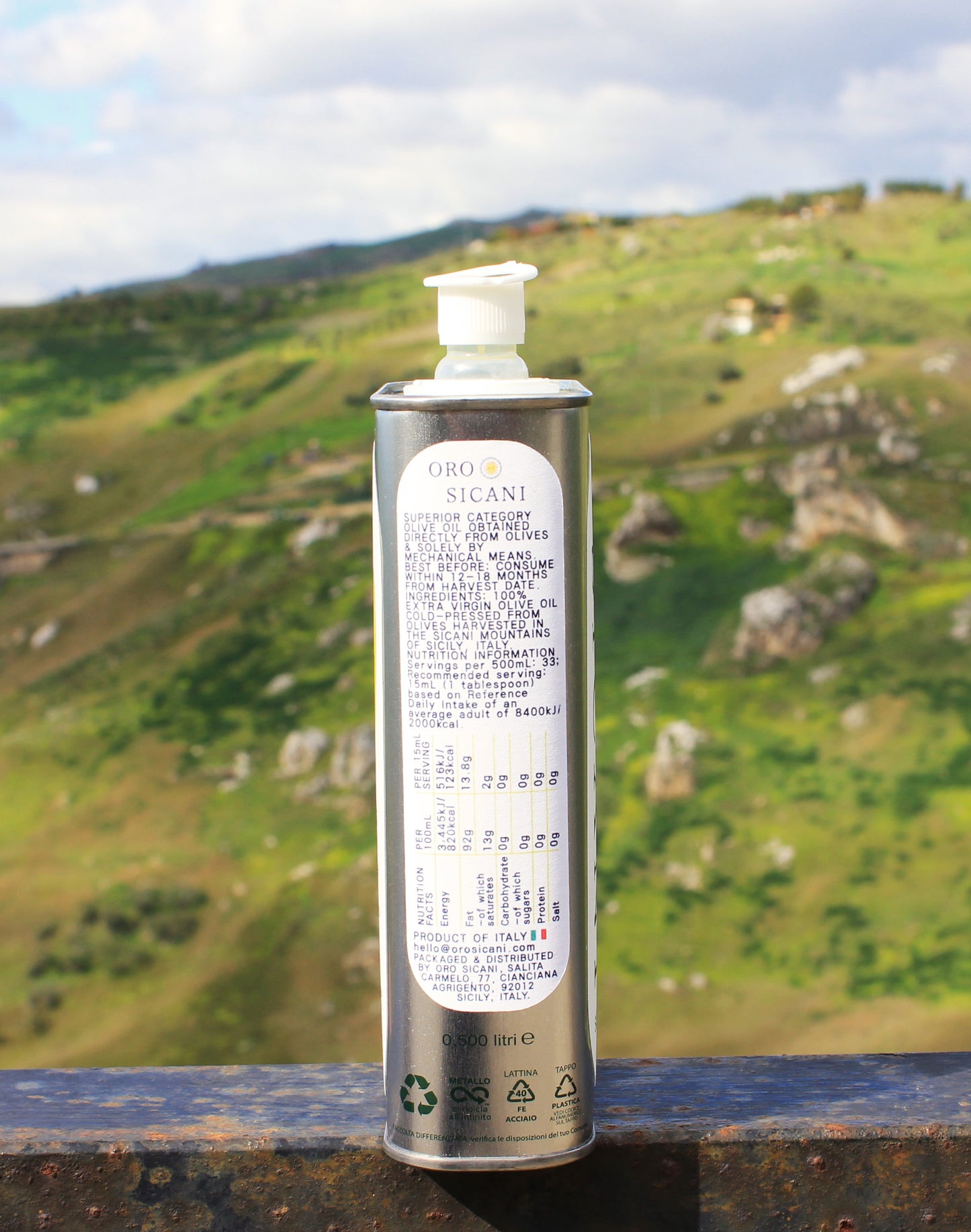 Close-up side profile of nutrition information of ORO SICANI Extra Virgin Olive Oil tin, placed on top of rusted railing with a lush, green mountain in the background.