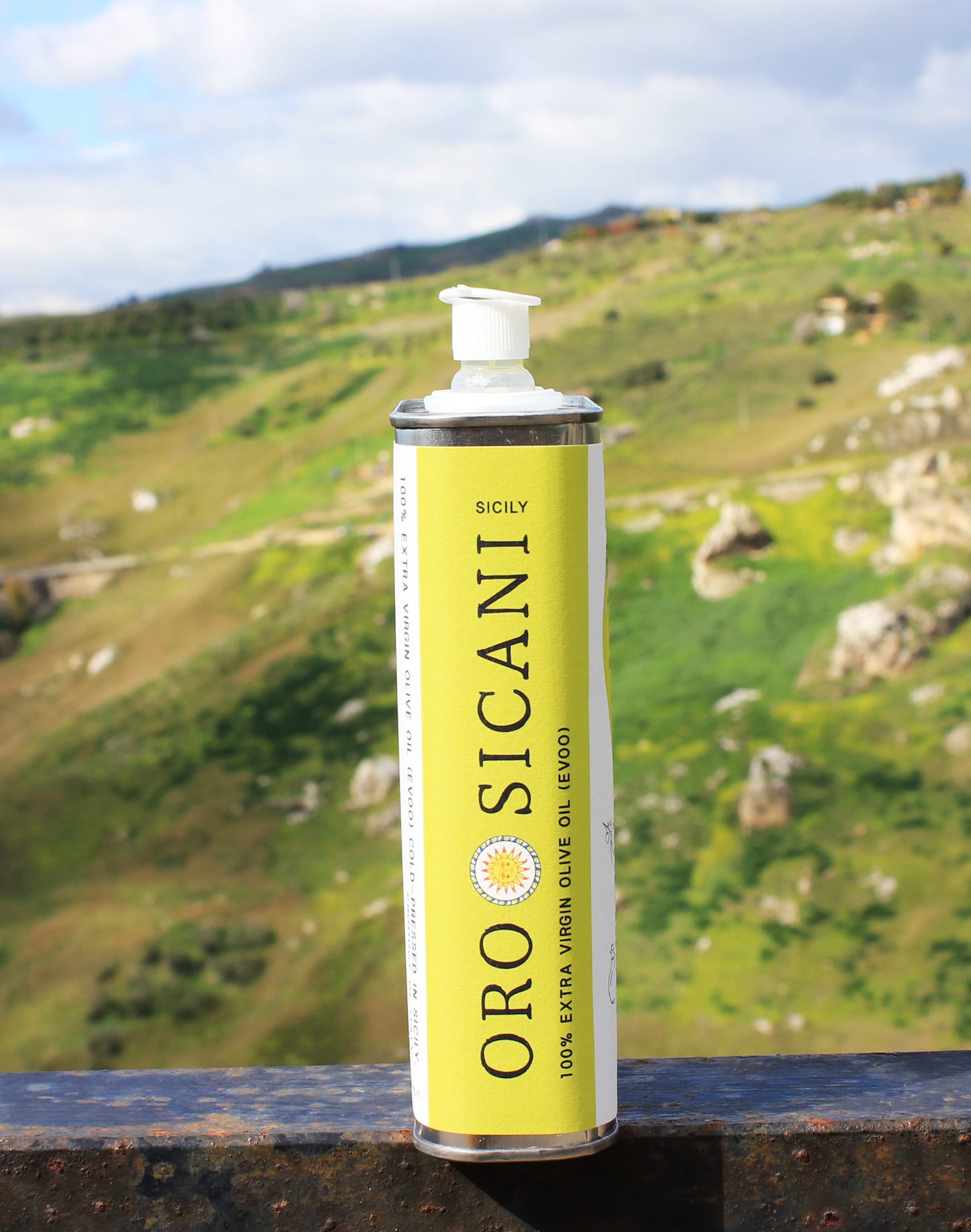 Close-up side profile of ORO SICANI Extra Virgin Olive Oil tin, placed on top of rusted railing with a lush, green mountain in the background.
