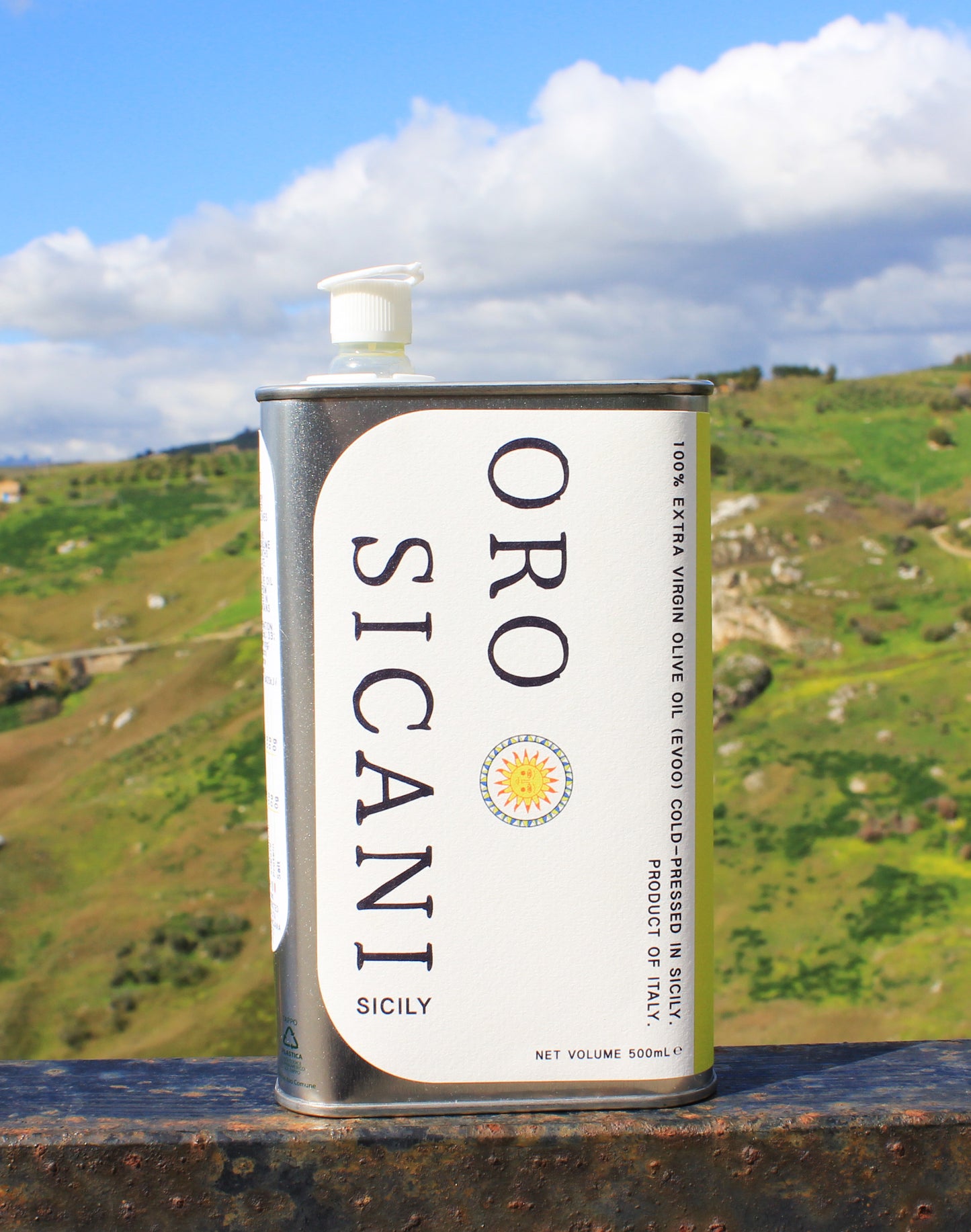Close-up of ORO SICANI Extra Virgin Olive Oil tin, placed on top of rusted railing with a lush, green mountain in the background.