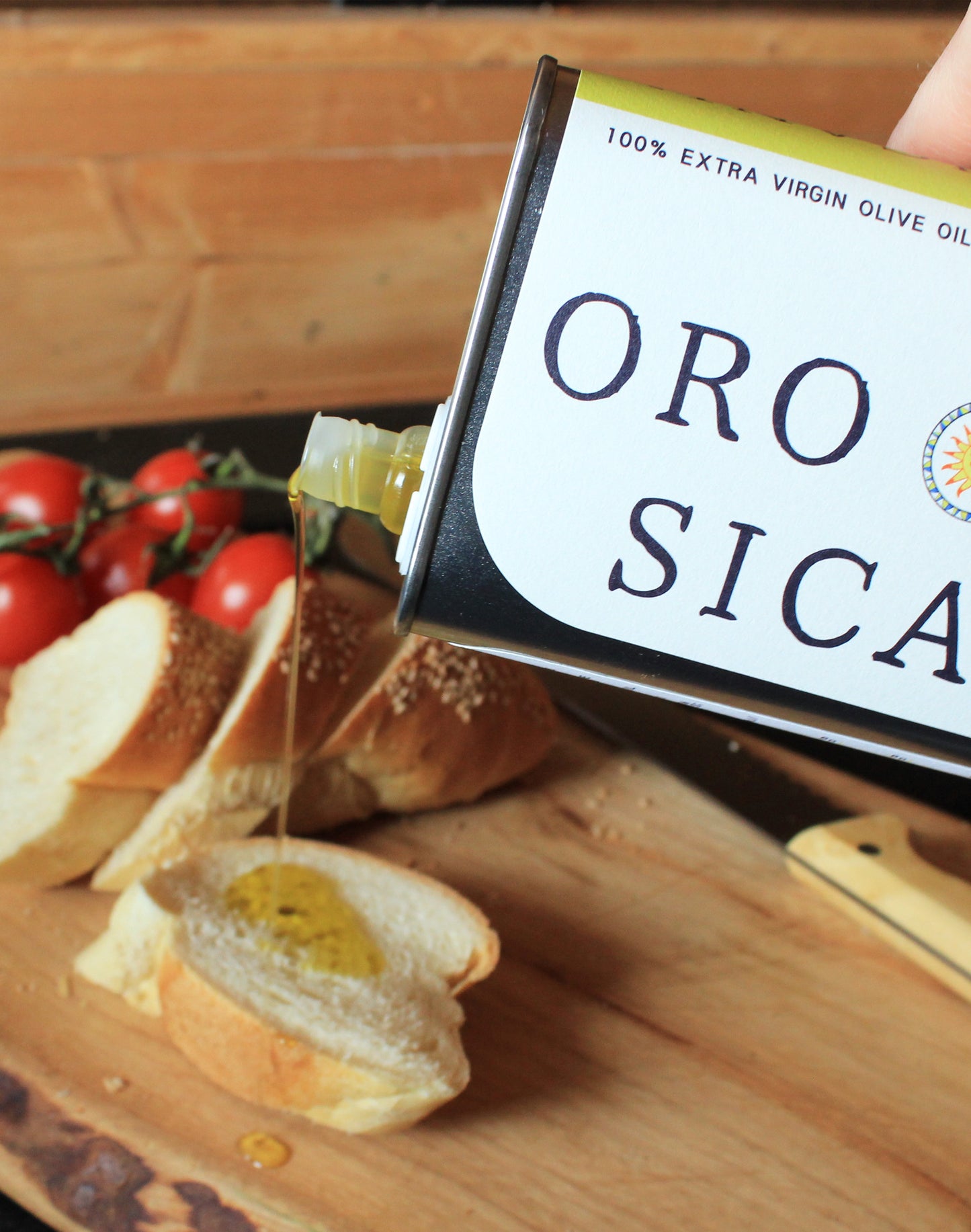 Close-up of ORO SICANI Extra Virgin Olive Oil being poured over a baguette slices on wooden chopping board with vine tomatoes.