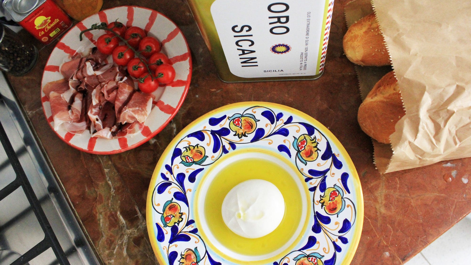 bird's eye view of a rosso marble kitchen countertop with oro sicani extra virgin olive oil encircling a burrata in a hand-painted ceramic bowl made in sicily, italy, as well as two baguettes in a brown paper bag, and an italian hand=painted ceramic plate of tomatoes and prosciutto