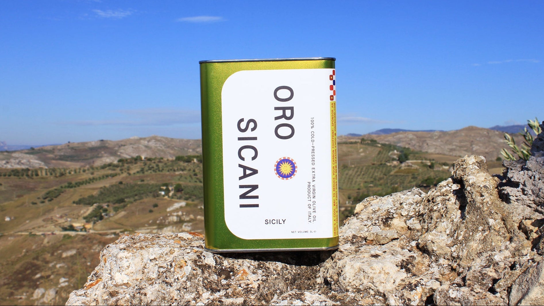 Oro Sicani extra virgin olive oil 3L tin on a rocky outcrop in the Sicani Mountains of Sicily, Italy, with a scenic background of rolling hills and olive fields in the distance