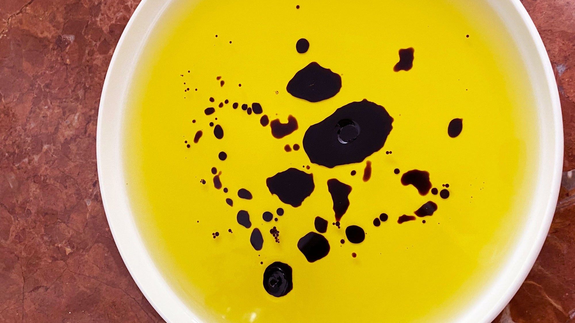 close up of oro sicani extra virgin olive oil with balsamic vinegar dotted throughout, on rosso marble kitchen countertop 