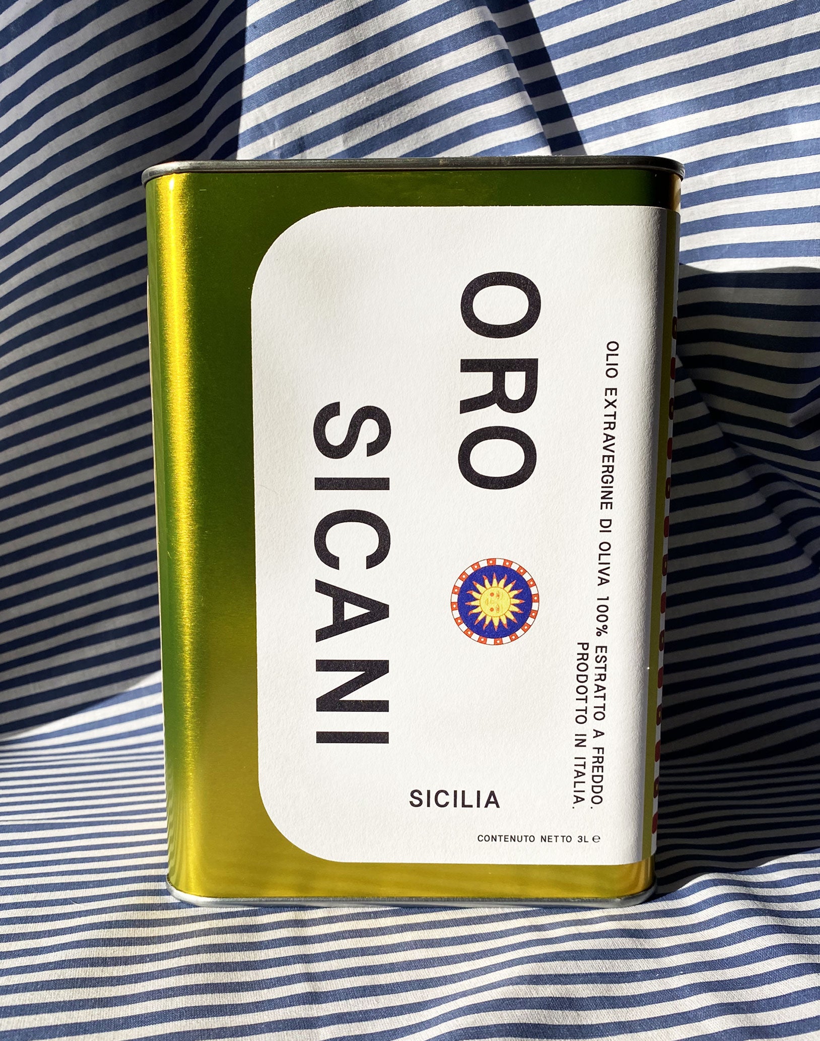 Close up of gold oro sicani extra virgin olive oil 3L tin with white label in front of blue-and-white-striped tablecloth backdrop.