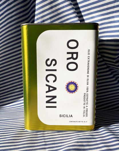 Close up of gold oro sicani extra virgin olive oil 3L tin with white label in front of blue-and-white-striped tablecloth backdrop.