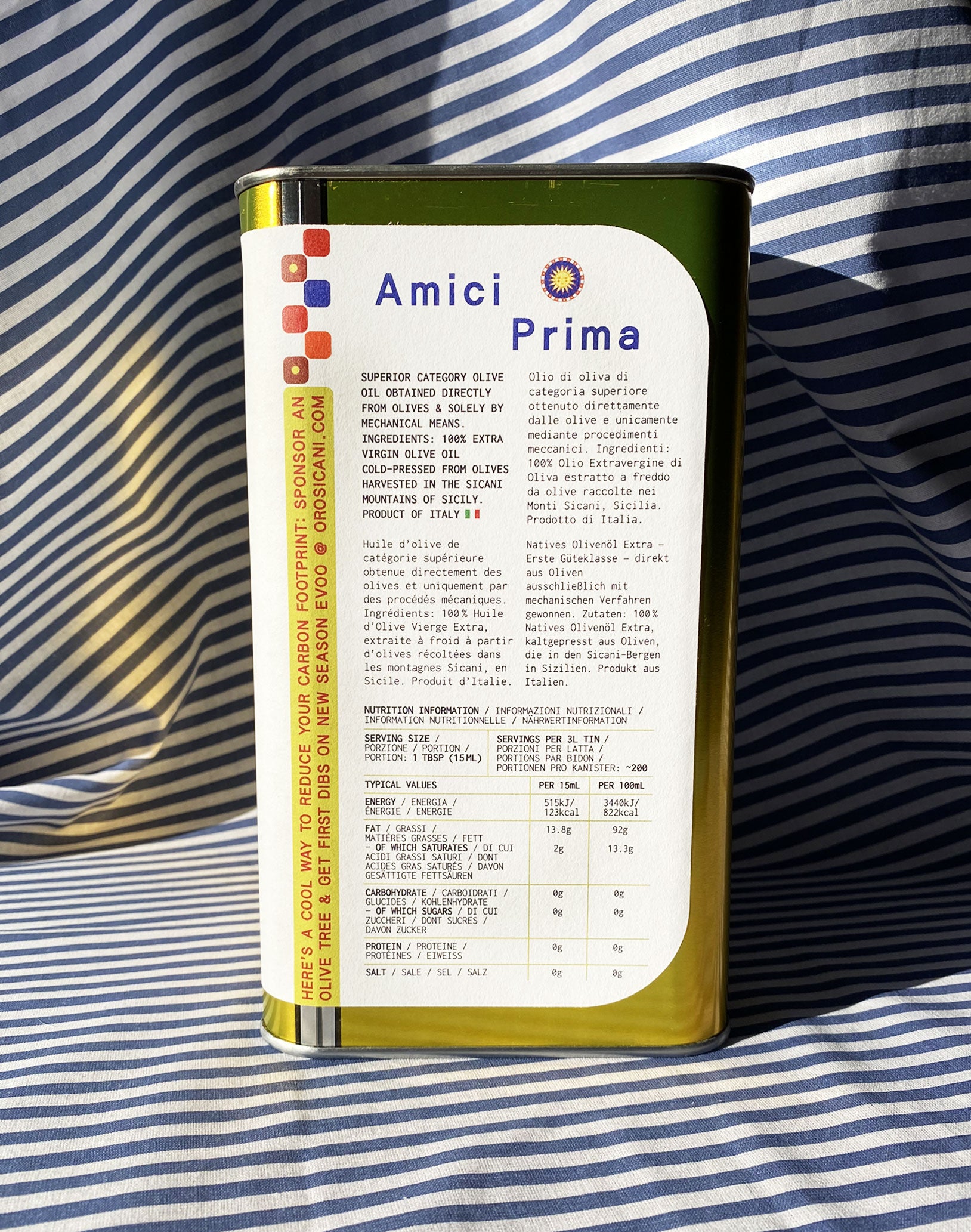 Close up of gold oro sicani extra virgin olive oil 3L tin with white nutrition label in front of blue-and-white-striped tablecloth backdrop.