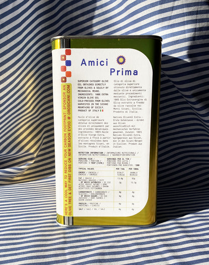 Close up of gold oro sicani extra virgin olive oil 3L tin with white nutrition label in front of blue-and-white-striped tablecloth backdrop.