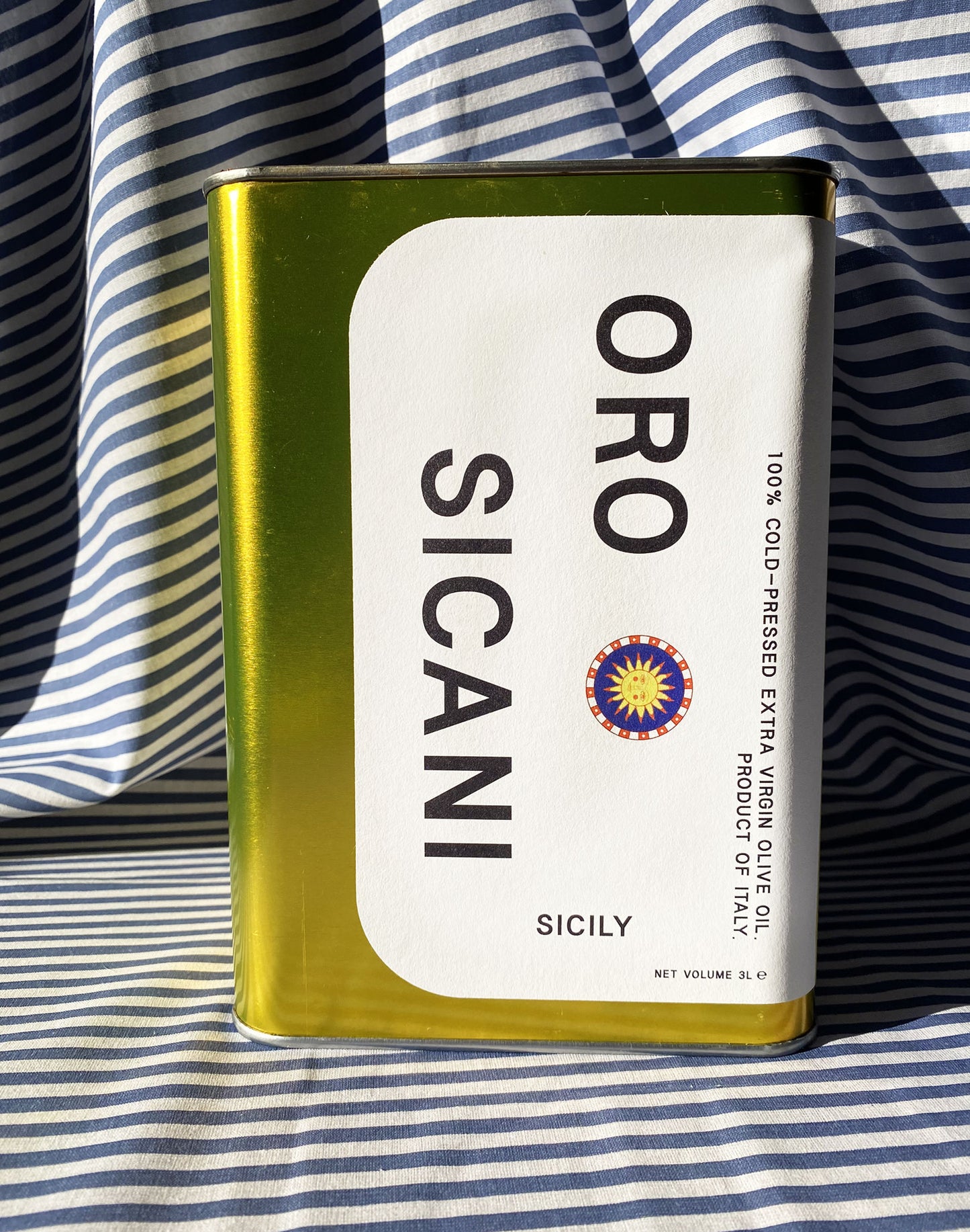 Close up of gold oro sicani extra virgin olive oil 3L tin with white label in front of blue-and-white-striped tablecloth backdrop.