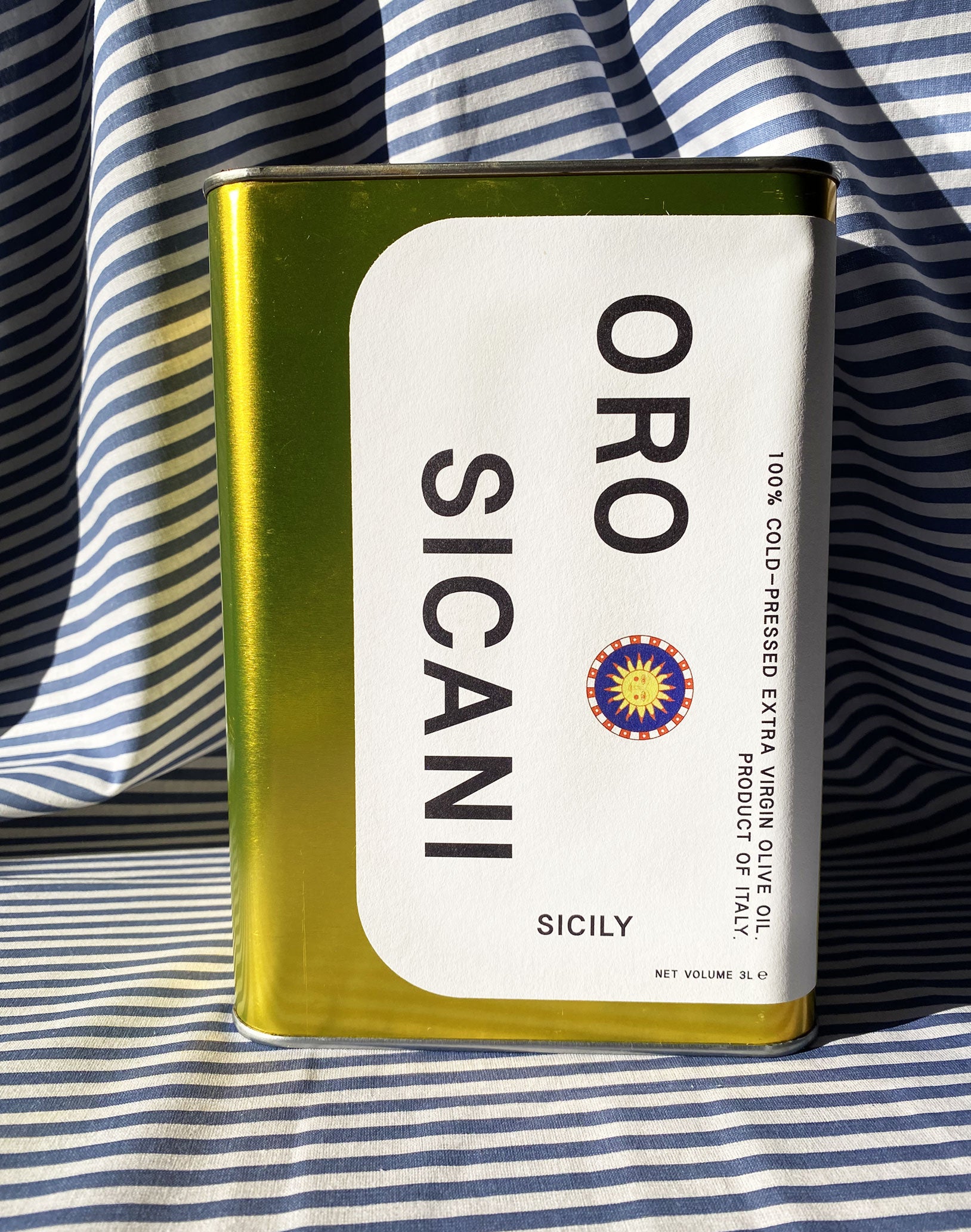 Close up of gold oro sicani extra virgin olive oil 3L tin with white label in front of blue-and-white-striped tablecloth backdrop.