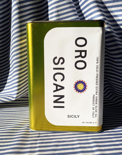 Close up of gold oro sicani extra virgin olive oil 3L tin with white label in front of blue-and-white-striped tablecloth backdrop.