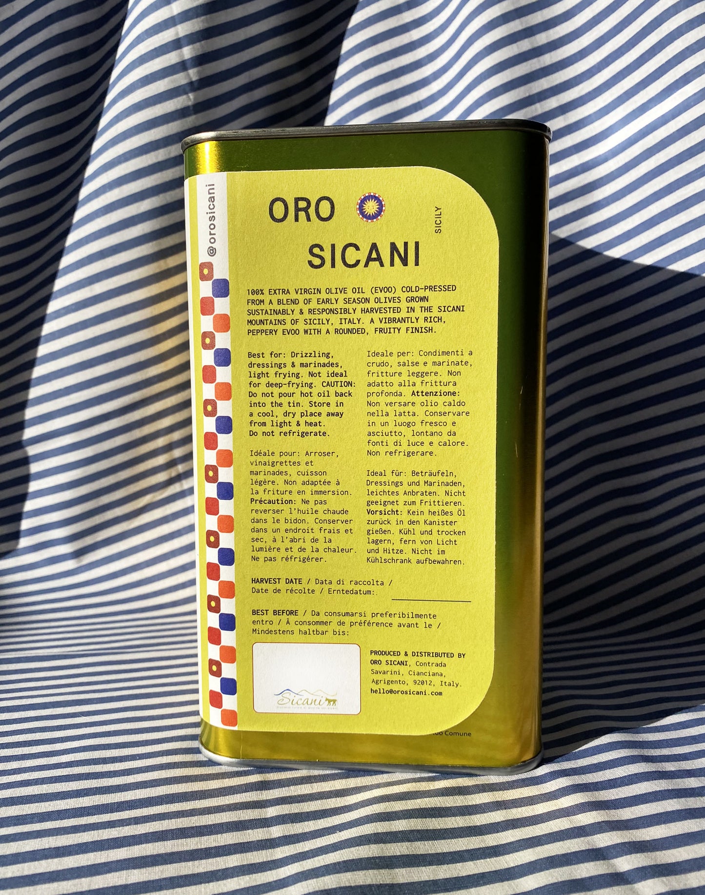 Close up of gold oro sicani extra virgin olive oil 3L tin with green information label in front of blue-and-white-striped tablecloth backdrop