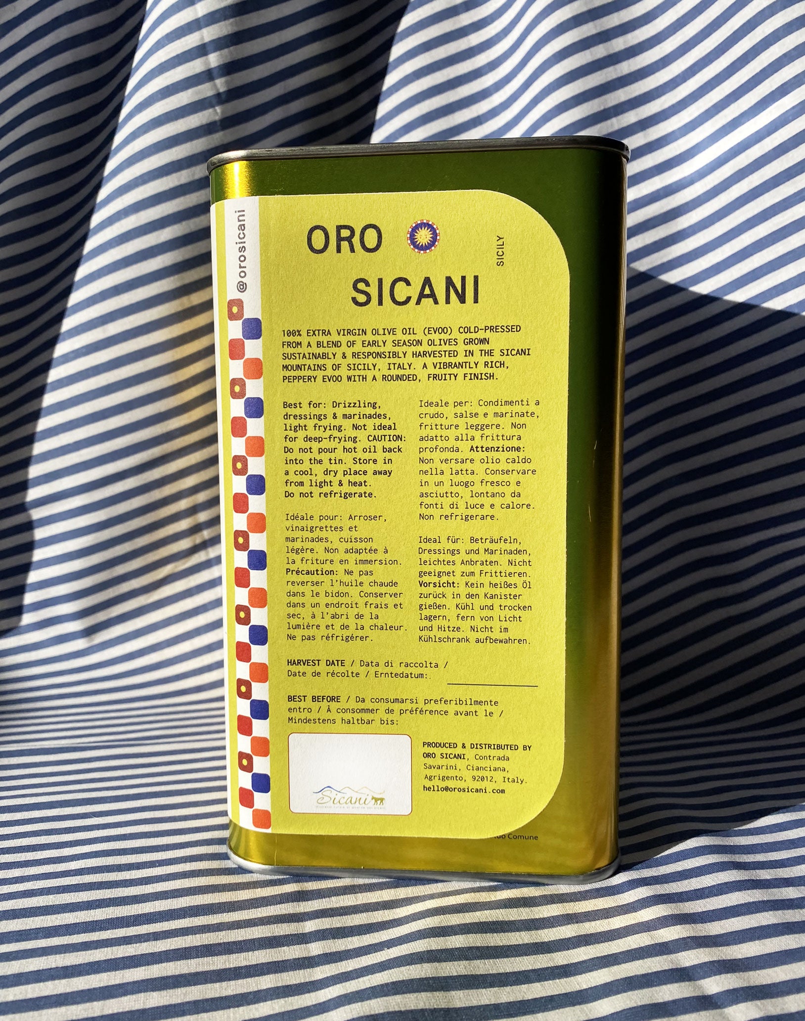 Close up of gold oro sicani extra virgin olive oil 3L tin with green information label in front of blue-and-white-striped tablecloth backdrop