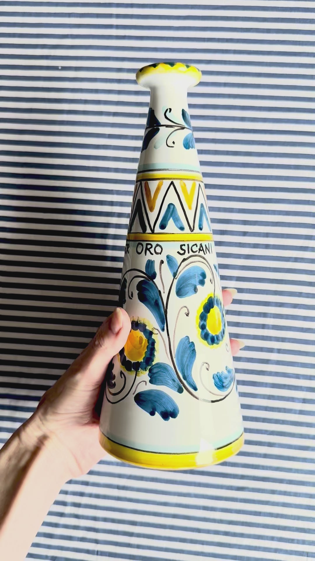 360 degree view of hand-painted Sicilian ceramic bottle with blue flower patterns in front of a striped background