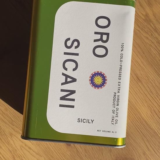 a bird's eye view video of gold oro sicani extra virgin olive oil 3l tin laying flat on a wooden table and shimmering in the sunlight. the evoo tin is being slowly turned so you see all four sides of the tin.