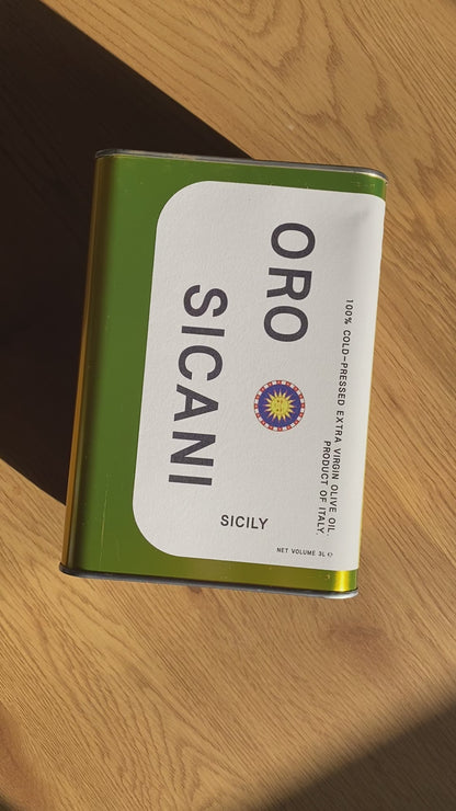 a bird's eye view video of gold oro sicani extra virgin olive oil 3l tin laying flat on a wooden table and shimmering in the sunlight. the evoo tin is being slowly turned so you see all four sides of the tin.